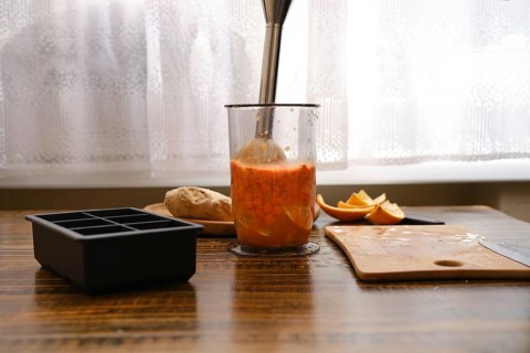 Mixing sea buckthorn and orange in a jar with immersion blender on a wooden table near the window .