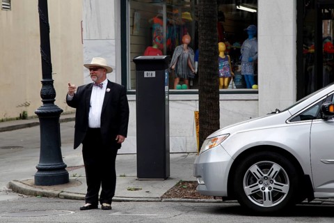 Elderly man in formal clothes stands in front of a shop window on a street corner. Modern car