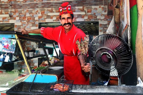 Smiling chef in red outfit offers brochettes at a barbecue area in Moulay Idris, Moulay Idris,