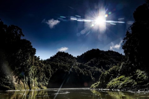 The sun shines over the wooded hills of the Whanganui River and meets calm water, zero
