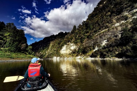 Person in a canoe paddles along steep, wooded cliffs under a dramatic sky, zero