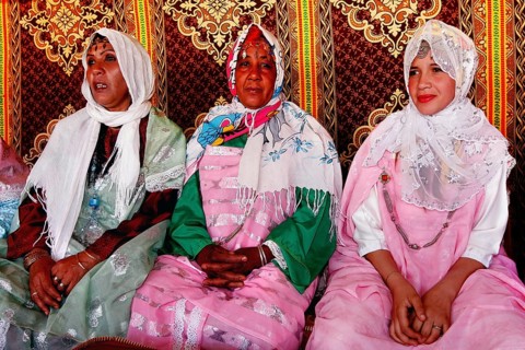 Three woman in traditional dress sit in a tent during the rose festival in El Kelâa m'Gouna, El