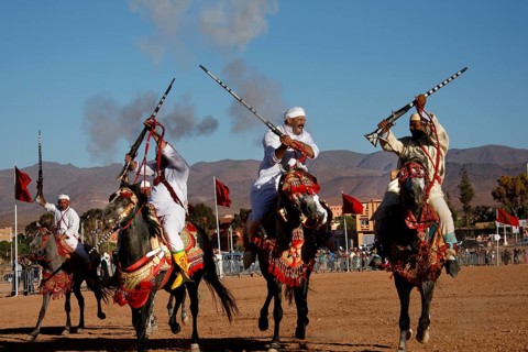 Three horsemen pass each other with rifles in traditional clothing, surrounded by dusty landscape,