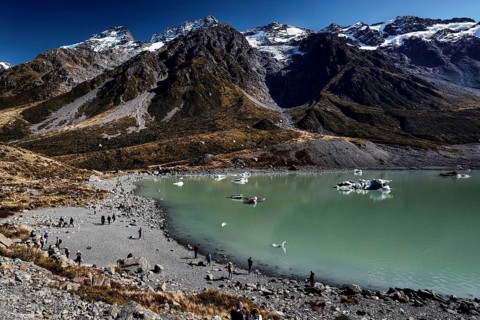 An expansive glacial lake surrounded by rugged mountains under clear skies, Mount Cook, New Zealand