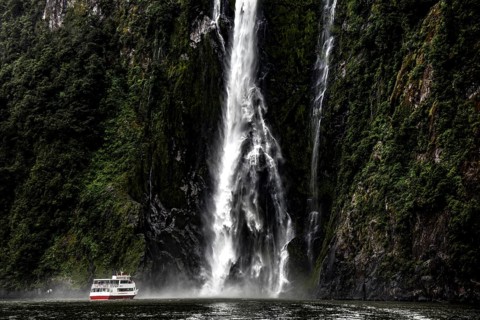 A mighty waterfall falls into the fjord, a boat glides past, surrounded by steep, green rocks, zero