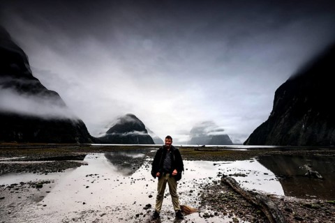Man standing in front of a dramatic sky on the shore of a fjord with reflecting water, Milford