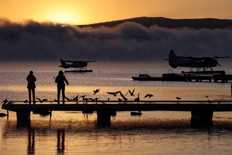 A seaplane on Lake Rotorua at sunrise, with silhouettes of people and birds on the dock, Rotorua,