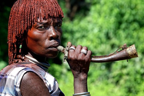 Hamer woman blowing a traditional horn surrounded by green vegetation, Turmi, Ethiopia