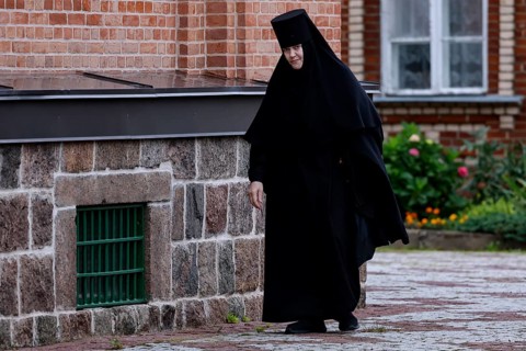 Nun in black clothes walks along the stone wall of the church, Kuremäe, Pühtitsa, Estonia