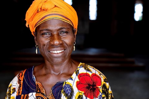 Woman in colorful dress with yellow headscarf smiles at the camera. Dark background, Bouaké,