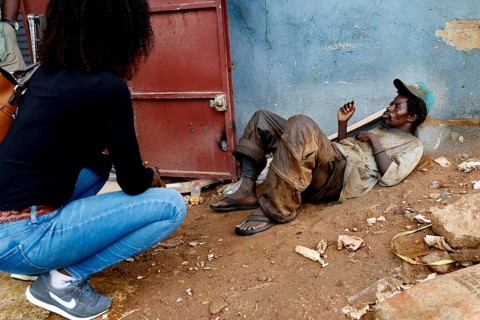 Mentally ill homeless person sits in front of a door. A woman approaches him on a dusty road,