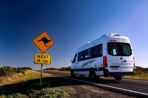 A motorhome drives along the Great Ocean Road past a kangaroo warning sign under clear blue skies,