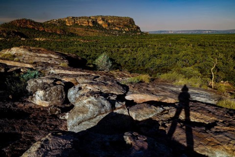 Shadows of a person on a rocky elevation in front of forest and rocks under clear sky, Nawurlandia,