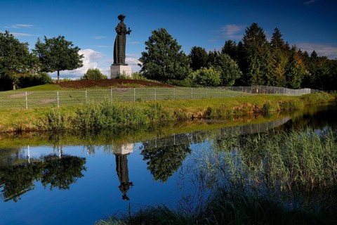 Socialist statue in Grutas Park is reflected in a calm body of water surrounded by trees,