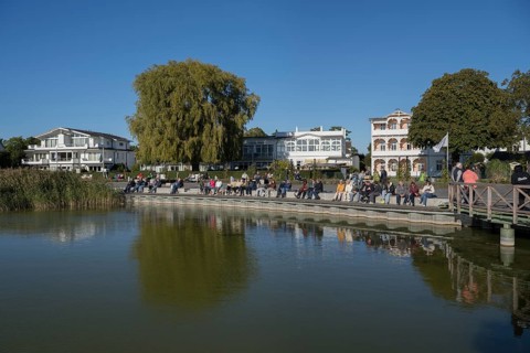 Tourists sunbathe on the promenade of Schmachter See, in the back buildings of spa architecture,