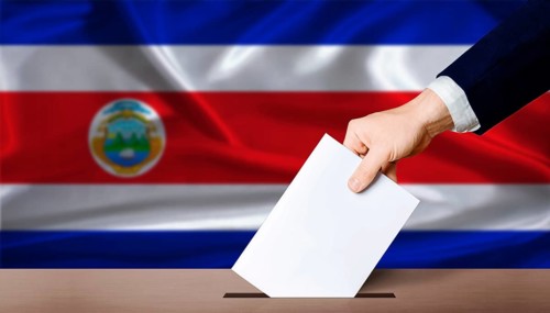 Hand placing a ballot in ballot box with the Costa Rican flag in the background. Costa Rica