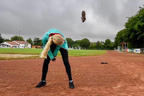 A woman on a sports field throws a rubber boot into the air under a cloudy sky, wellies long throw