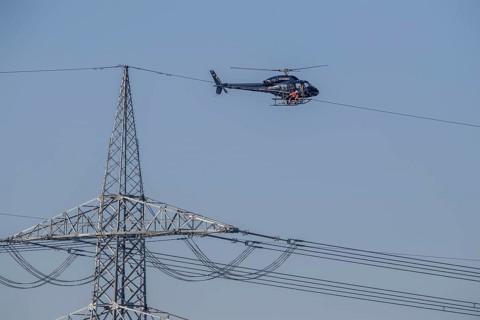A helicopter carries out technical work on power lines next to a mast under a blue sky, an employee