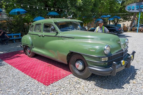Green vintage car standing on outdoor red carpet surrounded by colorful umbrellas and gravel floor,