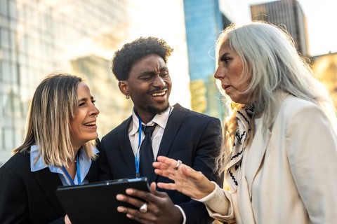 Diverse business professionals meet outdoors in the city, smiling and collaborating over a tablet