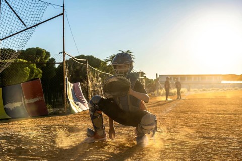 Boy catcher wearing protective gear and mitt, squatting on a dusty baseball field with golden
