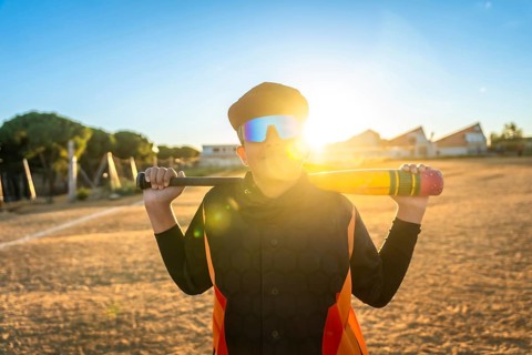 Youth baseball player in sunglasses and orange black jersey gripping a bat on a dusty field at