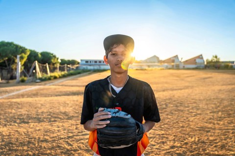 Young baseball player stands on a dusty field at golden hour, wearing a black uniform and cap,