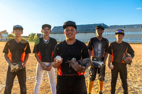 Group of diverse teenage baseball players standing on a dusty field, one holding a ball and glove,