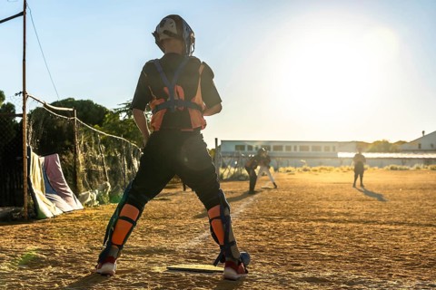 Catcher in full gear stands on a dusty baseball field at sunset, silhouetted against golden light