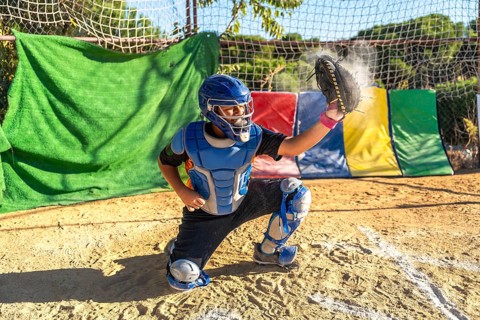 Boy catcher wearing helmet, chest protector, and leg guards, catching a baseball with glove,