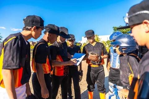 Baseball coach in black and orange uniform reviews strategy on a clipboard with attentive youth