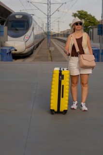 Adult female traveler standing on a railway platform with a yellow suitcase, looking around while