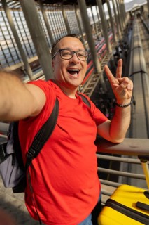Middle aged man wearing reading glasses smiles with teeth while taking a selfie and making a peace