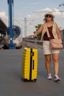 Adult woman walking alone on a city train platform, pulling a yellow suitcase and looking ahead