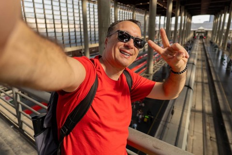 Middle aged man wearing sunglasses takes a cheerful selfie at a busy train station. Modern travel,
