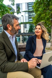 Two professional business colleagues, a man and a woman, are sitting on a bench in a city