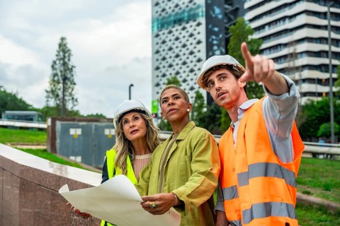 Diverse construction team in hard hats and safety vests reviews blueprints and points toward an