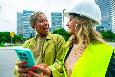 Two diverse colleagues. One an architect wearing a hardhat and safety vest. Looking at a smartphone