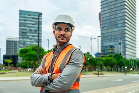 Young man wearing a hard hat and safety vest standing confidently with arms crossed in a cityscape,