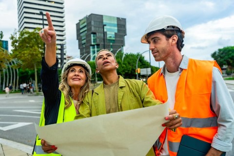 Diverse team of architects and engineers wearing hard hats and safety vests, examining blueprints