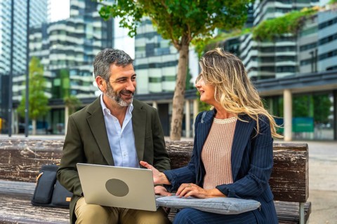 Two smiling business colleagues having an informal outdoor meeting on a bench, collaborating on a