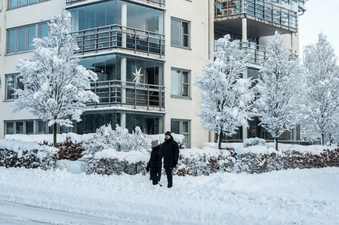 A couple walks on a snow-covered street in front of apartment buildings and snow-laden trees in