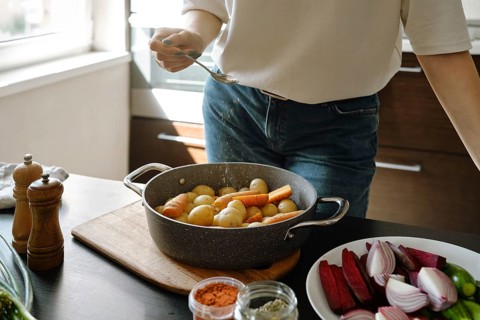 Unrecognizable woman sprinkles dried oregano on baby potatoes and carrots in a baking pan