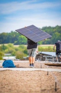 A man carries a solar panel on a construction site with a wooded background, new park and ride