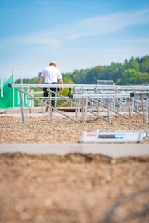 A worker assembles solar panel structures on a gravel roof under a blue sky, new park and ride