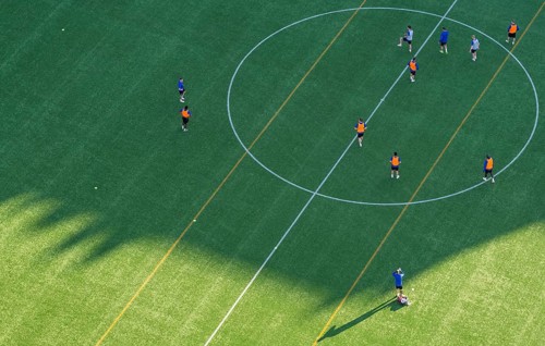 Players wearing colorful jerseys play soccer on a grass field from a bird's eye view from the tower