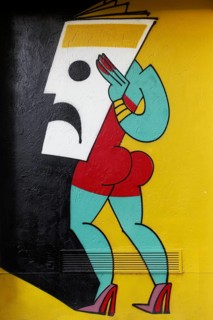 Stylized female figure holding mask in front of her face, colored mural by street art artist Mina
