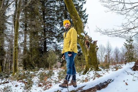 Man wearing a yellow jacket and beanie standing on a fallen tree trunk in a snowy forest, enjoying