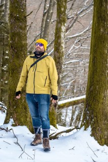 Man wearing a yellow winter jacket, jeans, and hiking boots exploring a snowy forest landscape,