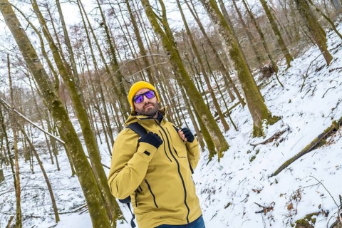 Man wearing a yellow beanie and jacket, sunglasses, and backpack, standing confident and looking at